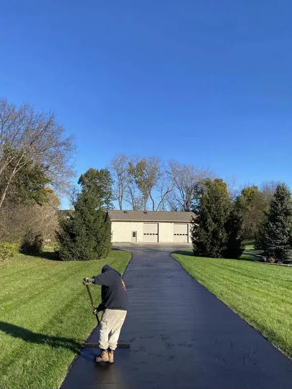 A worker applying sealcoat by squeegee along a long asphalt driveway