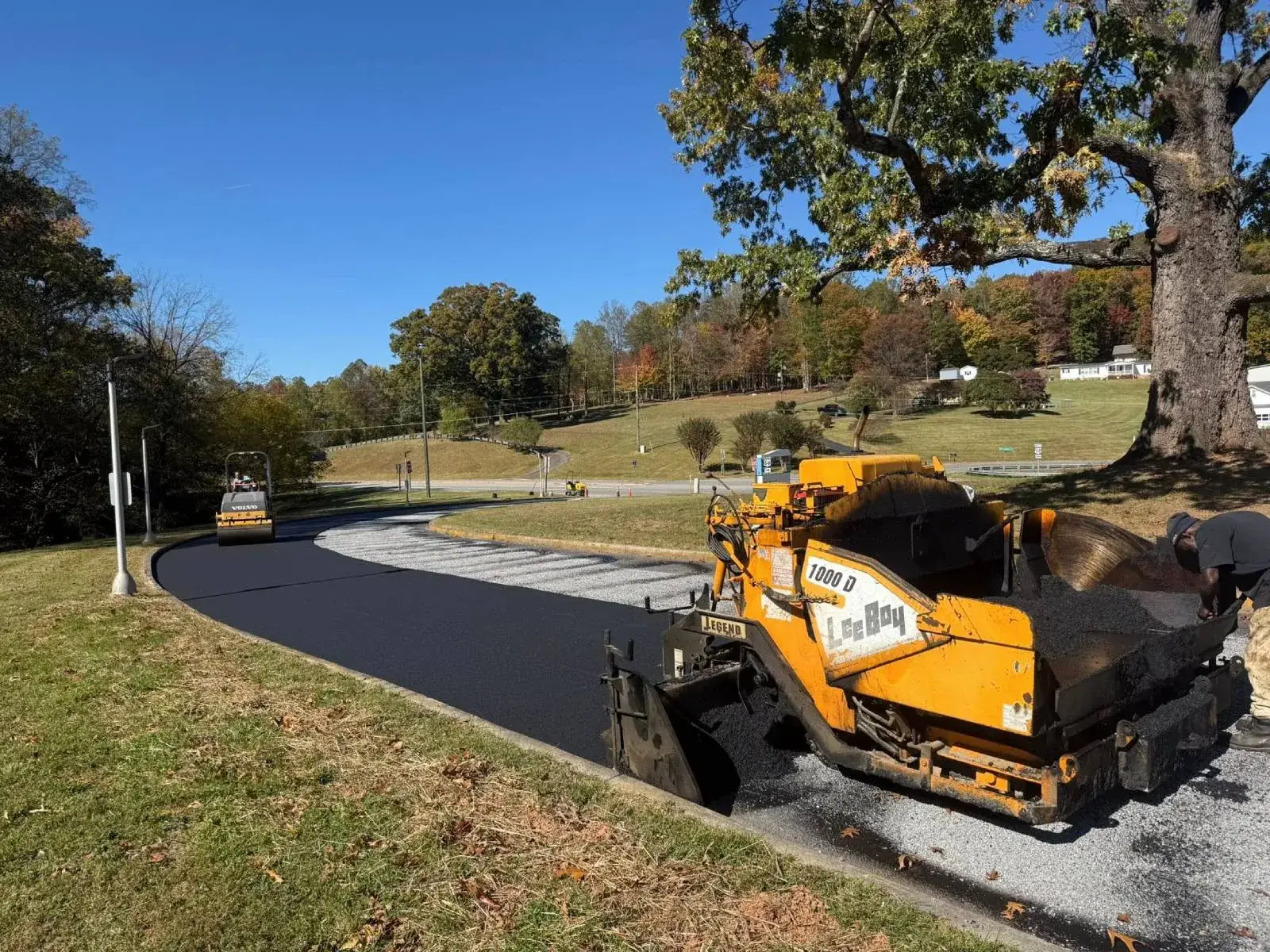 An asphalt paver laying a fresh mat of hot mix along a tree-lined road