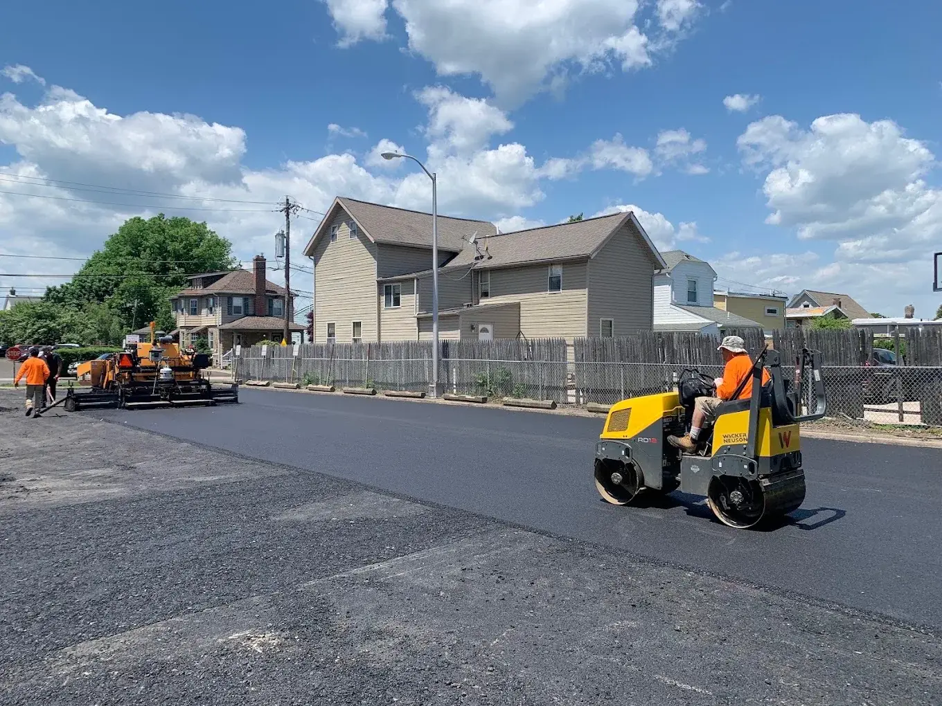 A paving crew laying a fresh asphalt surface across a residential street, roller in the foreground