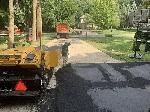 A paver laying new asphalt across a residential driveway beside a basketball hoop