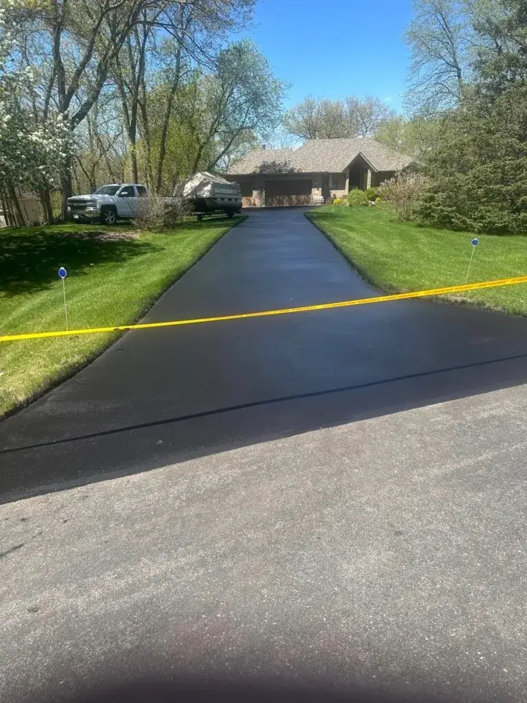 A long, freshly sealed asphalt driveway running up to a ranch house with a green lawn either side